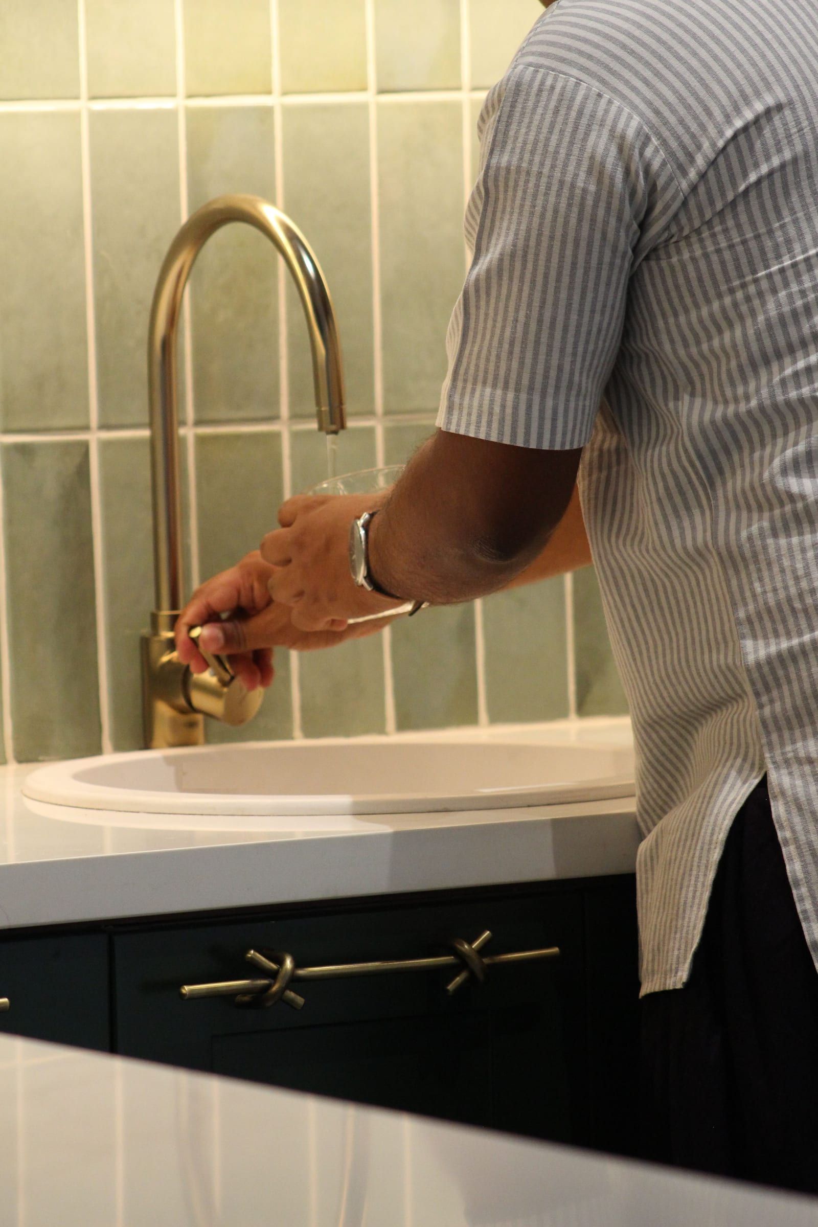 Hands at an arched brass tap over a round sink, a brass knot handle on the green cabinet below